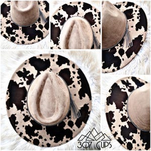 May include: A beige fedora-style hat with a cowhide print brim and a matching tassel. The hat features a neutral-toned crown and a black and white spotted pattern on the brim. The hat is a stylish accessory.