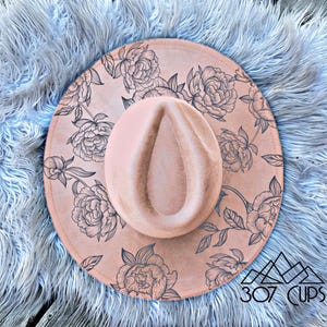May include: A blush-colored fedora hat with a floral design. The hat features a wide brim and a center crease. The floral pattern is in black ink. The hat is on a light blue fluffy surface. The text "307 CUPS" is visible.