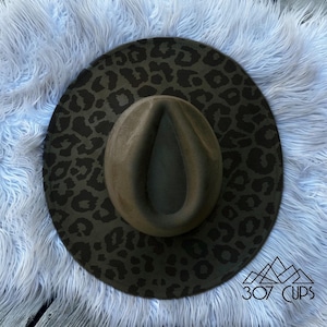May include: A fedora-style hat with a wide brim featuring a dark green leopard print. The crown of the hat is a solid olive green. The hat is displayed on a light blue faux fur surface.