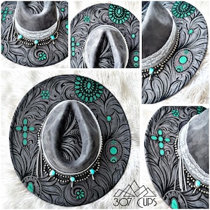 Może przedstawiać: Szary kapelusz kowbojski z dekoracyjnym wzorem kwiatowym w kolorze czarnym i turkusowym. Kapelusz posiada szary rondo, białą koronkową opaskę i turkusowe zdobienia. Kapelusz ma wzór krzyża i srebrny naszyjnik z koralików.