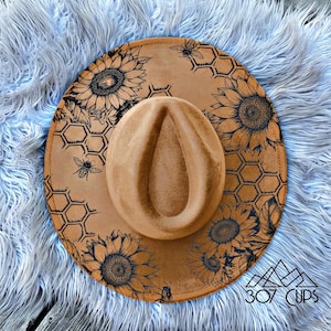 Op de afbeelding: Een bruin fedora hoed met een brede rand, versierd met zonnebloemen en honingraatpatronen. De hoed heeft een druppelvormige kroon en is versierd met zwarte illustraties van zonnebloemen, bijen en honingraatpatronen. De tekst "307 CUPS" is zichtbaar.