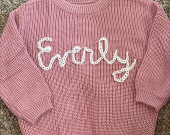 Puede incluir: Un suéter rosa tejido con el nombre "Everly" bordado en blanco en cursiva en el pecho. El suéter tiene una textura acanalada, mangas largas y cuello redondo. La prenda es para niños.