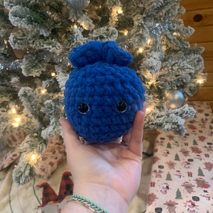 Puede incluir: Un juguete de peluche azul de crochet con forma de ar&aacute;ndano con ojos negros. El juguete est&aacute; siendo sostenido en una mano con una pulsera verde y dorada.