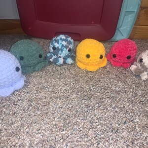Puede incluir: Seis peluches de pulpo de crochet en diferentes colores, incluyendo morado, verde, azul, amarillo, rojo y gris. Los peluches tienen ojos negros y est&aacute;n dispuestos en una fila sobre una alfombra.