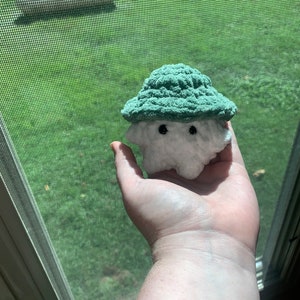 Puede incluir: Un juguete de peluche blanco tejido a crochet con un sombrero tejido a crochet verde. El juguete tiene dos ojos negros y una peque&ntilde;a boca.