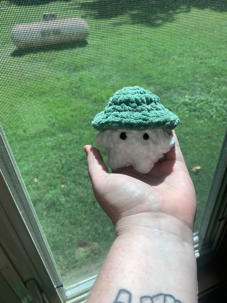 Puede incluir: Un juguete de peluche blanco tejido a crochet con un sombrero tejido a crochet verde. El juguete tiene dos ojos negros y una peque&ntilde;a boca.