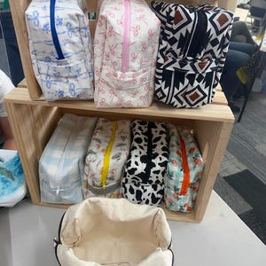 May include: A collection of rectangular fabric pouches with zippered closures, featuring various patterns including blue ribbons, pink bows, geometric shapes, light blue gingham, bees, cow print, and peaches, displayed on wooden shelves.