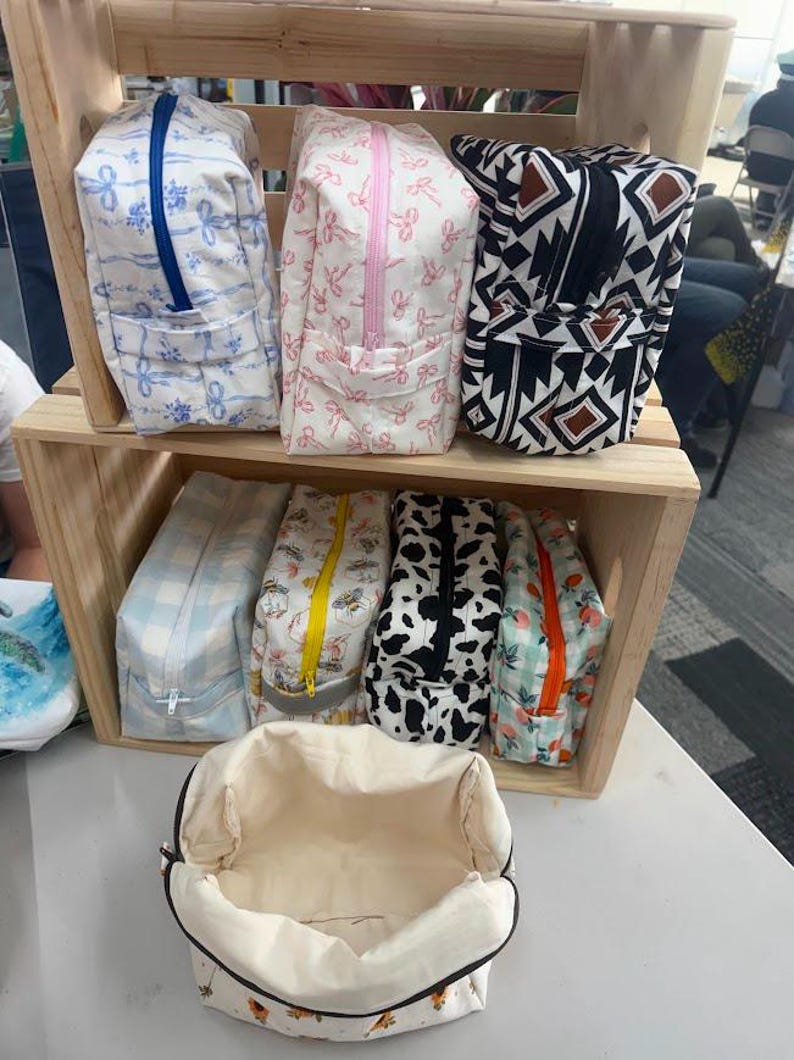 May include: A collection of rectangular fabric pouches with zippered closures, featuring various patterns including blue ribbons, pink bows, geometric shapes, light blue gingham, bees, cow print, and peaches, displayed on wooden shelves.
