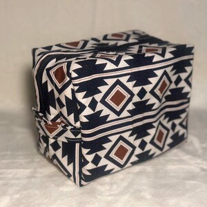 May include: A rectangular fabric storage bag featuring a repeating geometric pattern in black, white, and brown. The bag has a zippered top closure and a small fabric handle attached to the side panel, constructed with a structured boxy shape.