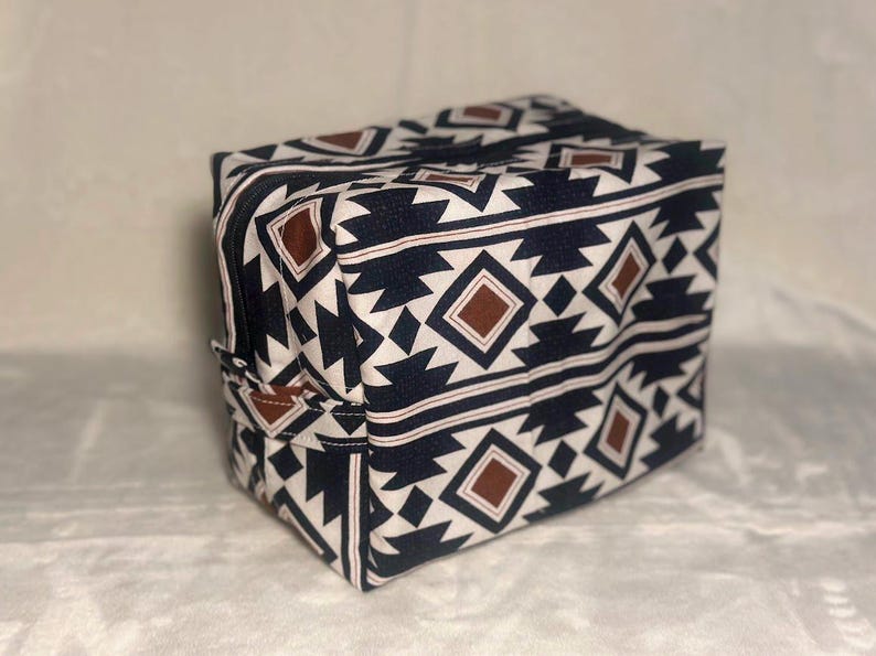 May include: A rectangular fabric storage bag featuring a repeating geometric pattern in black, white, and brown. The bag has a zippered top closure and a small fabric handle attached to the side panel, constructed with a structured boxy shape.