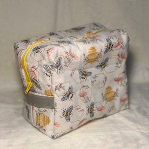 May include: A rectangular fabric pouch featuring a white background with a repeating pattern of bees, yellow beehives, and pink flowers. The bag includes a bright yellow zipper closure along the top and a solid gray fabric handle on the side.