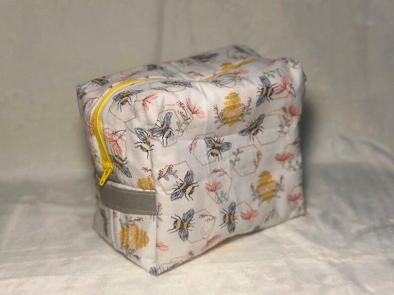 May include: A rectangular fabric pouch featuring a white background with a repeating pattern of bees, yellow beehives, and pink flowers. The bag includes a bright yellow zipper closure along the top and a solid gray fabric handle on the side.