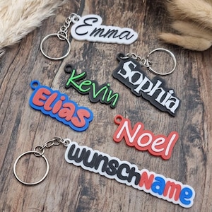 Könnte beinhalten: Eine Sammlung personalisierter Schlüsselanhänger mit Namen in verschiedenen Schriftarten und Farben, darunter Emma in Schwarz auf Weiß, Kevin in Grün, Sophia in Weiß auf Schwarz, Elias in Rot auf Blau, Noel in Weiß auf Rot und wunschName in Schwarz und Rot auf Weiß.