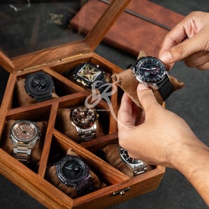 Peut inclure: Une boîte en bois pour montres avec un couvercle en verre, présentant plusieurs montres-bracelets. Une montre est manipulée, tandis que d'autres reposent sur des coussins bruns à l'intérieur de la boîte. Les montres présentent divers designs, dont des tons argentés et noirs.
