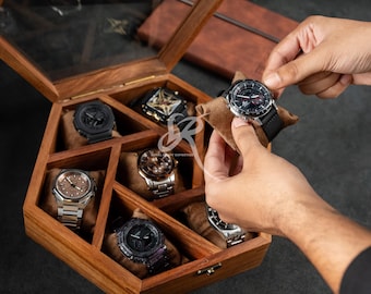 Puede incluir: Una caja de madera para relojes con tapa de cristal, que muestra varios relojes de pulsera. Se está manipulando un reloj, mientras que otros descansan sobre cojines marrones dentro de la caja. Los relojes presentan varios diseños, incluyendo tonos plateados y negros.