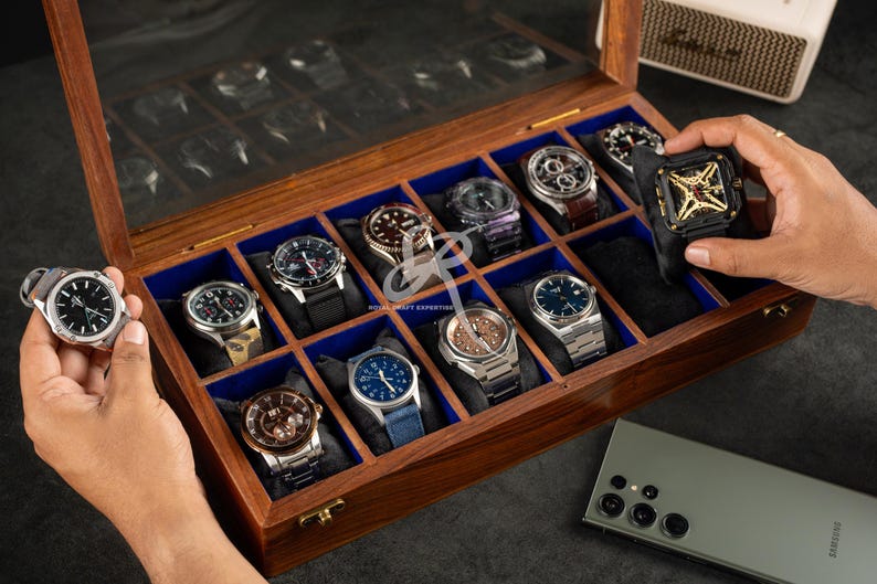 May include: A rectangular wooden watch display case with a glass lid, featuring twelve individual compartments lined with blue fabric. The case holds various wristwatches on black cushions, with a gold-tone metal latch on the front edge.