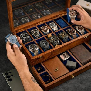 May include: A wooden watch storage box with a glass lid, featuring multiple compartments lined with blue velvet. The box contains various wristwatches, a brown leather wallet, a metal lighter, and a small metal accessory, organized in slots.
