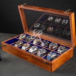 May include: A rectangular wooden watch display case with a hinged glass lid and gold-toned metal latches. The interior features twelve individual compartments lined with blue fabric, each containing a wristwatch on a cushion.