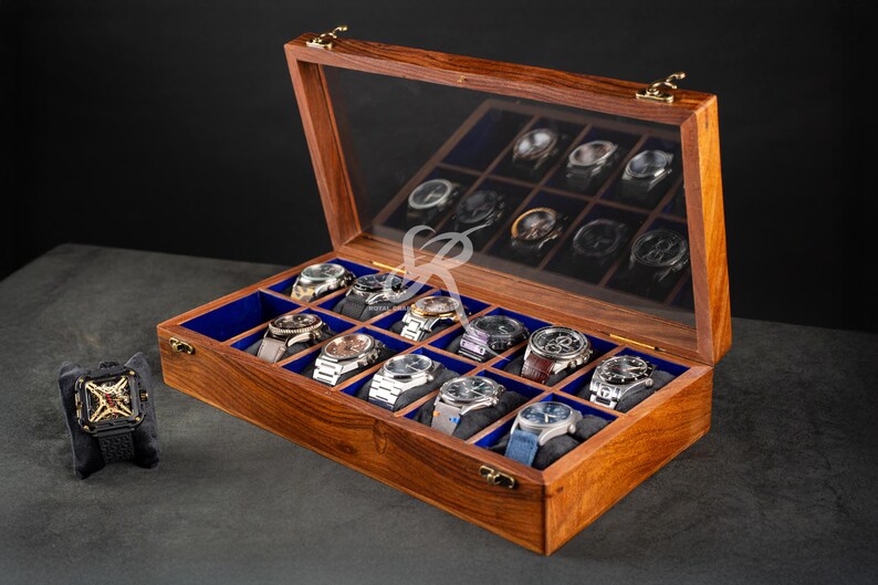 May include: A rectangular wooden watch display case with a hinged glass lid and gold-toned metal latches. The interior features twelve individual compartments lined with blue fabric, each containing a wristwatch on a cushion.