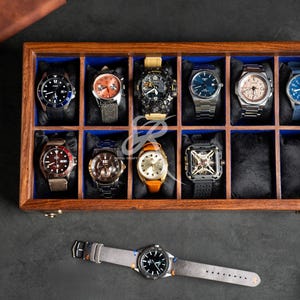 May include: A rectangular wooden watch display case with a glass lid, featuring twelve individual compartments lined with blue fabric. Eleven compartments contain various wristwatches with diverse dials and straps, while one is empty.