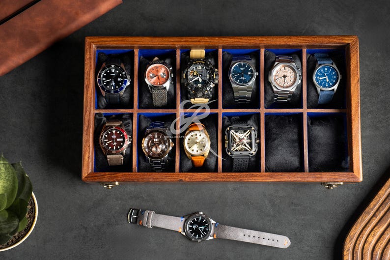 May include: A rectangular wooden watch display case with a glass lid, featuring twelve individual compartments lined with blue fabric. Eleven compartments contain various wristwatches with diverse dials and straps, while one is empty.