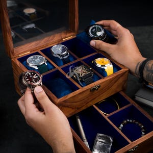 May include: A wooden watch box with a glass lid and blue velvet interior featuring multiple compartments. The box contains several wristwatches with various colored dials and straps, a metal pen, a lighter, and a beaded bracelet in the drawer.