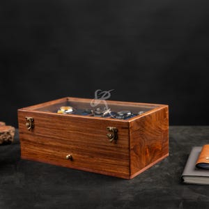 May include: A rectangular wooden watch storage box with a clear glass lid, featuring a rich brown wood grain finish, two brass-coloured metal latches on the front, and a small round knob for a drawer, containing multiple watches inside.