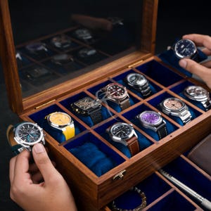 May include: A wooden watch display case with a glass lid and blue velvet interior, featuring multiple compartments holding various wristwatches with different colored dials and straps, including yellow, brown, and black bands.