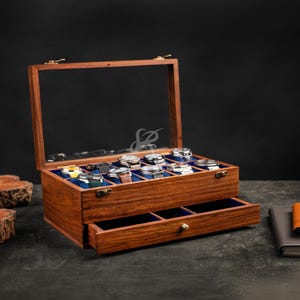 May include: A rectangular wooden watch display case with a hinged glass lid and a pull-out drawer. The interior features blue velvet lining with individual compartments for eight wristwatches, each resting on a small blue cushion.