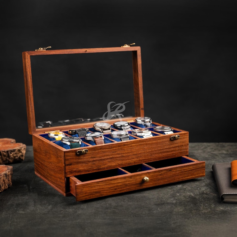 May include: A rectangular wooden watch display case with a hinged glass lid and a pull-out drawer. The interior features blue velvet lining with individual compartments for eight wristwatches, each resting on a small blue cushion.