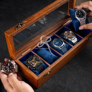 May include: A rectangular wooden watch display case with a clear glass lid and a brass latch. The interior features five individual compartments lined with blue velvet, each containing a soft blue cushion for holding a wristwatch securely.