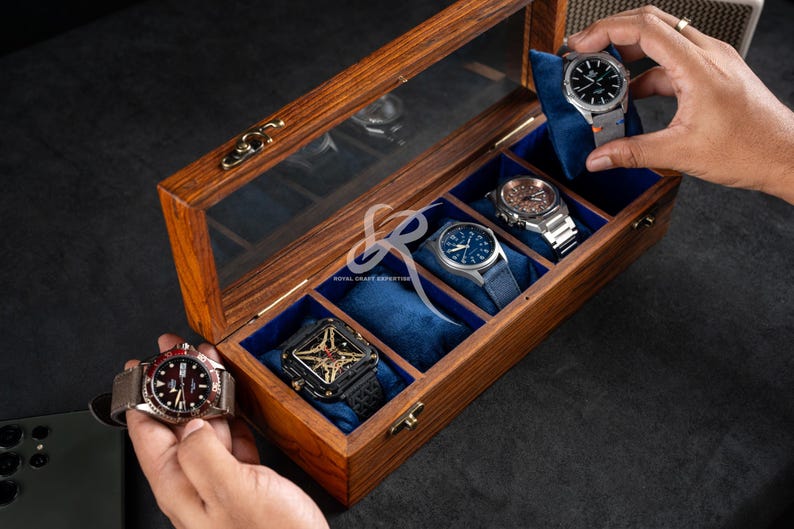 May include: A rectangular wooden watch display case with a clear glass lid and a brass latch. The interior features five individual compartments lined with blue velvet, each containing a soft blue cushion for holding a wristwatch securely.