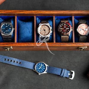 May include: A rectangular wooden watch box with a dark brown finish and blue velvet lining, containing four wristwatches on cushions. A fifth wristwatch with a blue canvas strap lies horizontally in front of the box. The box features gold-tone metal latches.