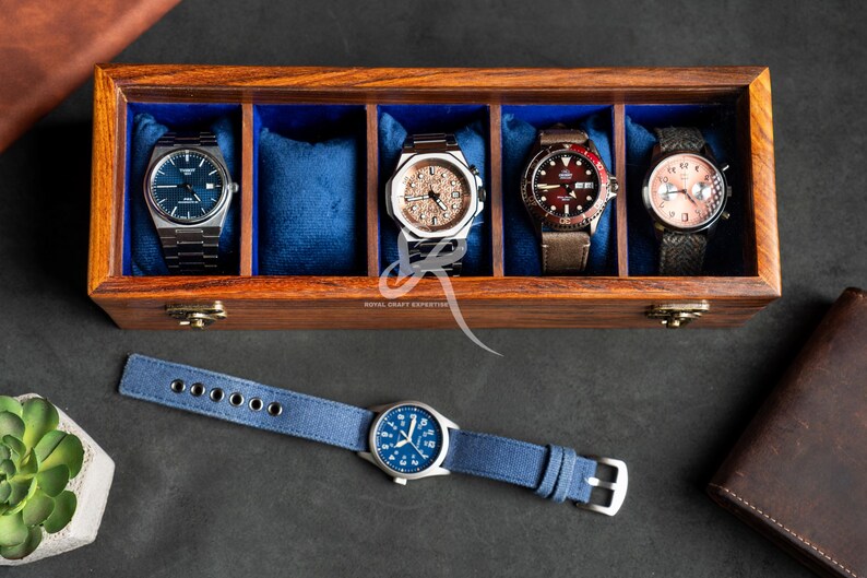 May include: A rectangular wooden watch box with a dark brown finish and blue velvet lining, containing four wristwatches on cushions. A fifth wristwatch with a blue canvas strap lies horizontally in front of the box. The box features gold-tone metal latches.