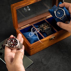 May include: A rectangular wooden watch box with a glass lid and blue velvet interior compartments. The box contains a square black watch, while two other watches with blue and red faces are held outside the box near a smartphone.