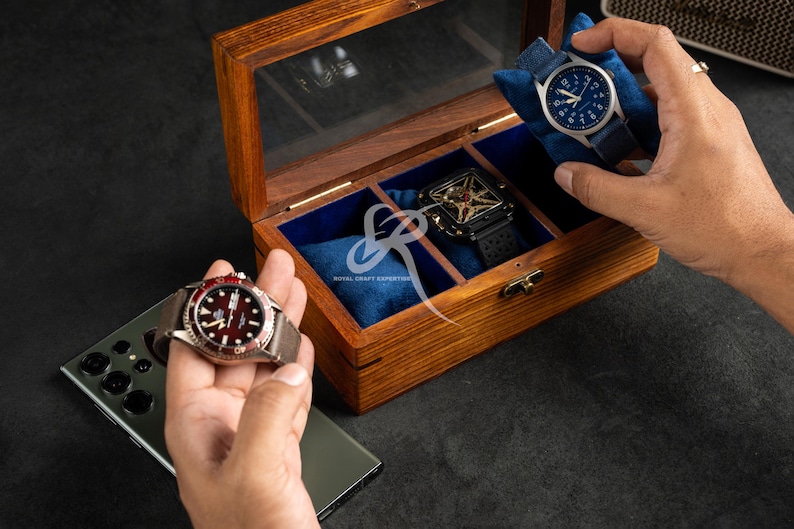 May include: A rectangular wooden watch box with a glass lid and blue velvet interior compartments. The box contains a square black watch, while two other watches with blue and red faces are held outside the box near a smartphone.