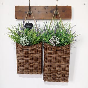 May include: Two brown wicker hanging baskets with faux greenery and lavender flowers. The baskets are hung on a wooden rack with hooks. A small black chalkboard sign with the word "welcome" is attached to one of the baskets.