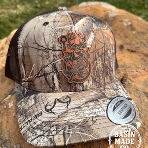 May include: Camouflage baseball cap with a brown mesh back and a leather patch featuring a deer, fish, and turkey. The hat has a curved brim and a snapback closure. The text "BASIN MADE CO." is visible.