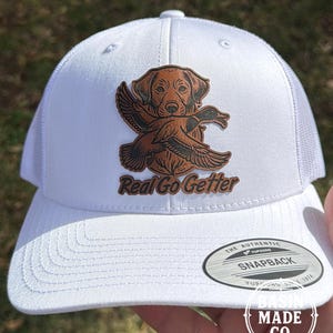 May include: White trucker hat with a brown leather patch featuring a dog and duck graphic. The text "Real Go Getter" is below the graphic. The hat has a curved brim and a snapback closure.