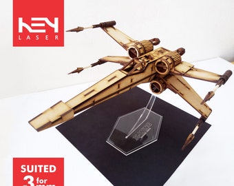 May include: A wooden X-Wing model with intricate laser-cut details, displayed on a clear stand. The model has a light brown color with darker brown accents. The stand is on a black square. The text "X-WING" is visible on the stand.