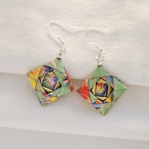 May include: A pair of handmade origami earrings. Each earring features a square, diamond-shaped design with a green border and colorful, patterned interior. The earrings have silver-colored fish hook ear wires. The earrings are displayed on a white surface.