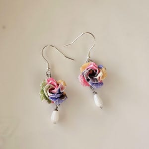 May include: A pair of floral earrings. Each earring has a fabric flower in pink, yellow, green, and purple, with a white teardrop bead hanging below. The earrings have silver hooks. The earrings are handmade.