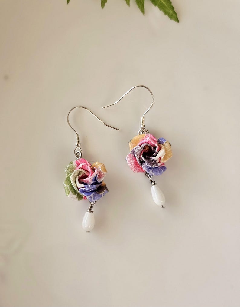 May include: A pair of floral earrings. Each earring has a fabric flower in pink, yellow, green, and purple, with a white teardrop bead hanging below. The earrings have silver hooks. The earrings are handmade.