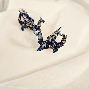 May include: A pair of origami crane earrings. The earrings are crafted from fabric featuring a navy blue, light blue, and gold design. The cranes are displayed on a white background.