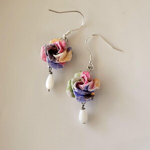 May include: Earrings featuring fabric flowers in pink, yellow, blue, and green. Each earring has a white, teardrop-shaped bead suspended beneath the flower. The earrings have silver-coloured hooks. A delicate and colourful accessory.