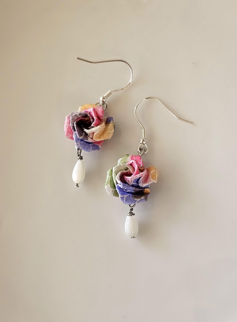May include: Earrings featuring fabric flowers in pink, yellow, blue, and green. Each earring has a white, teardrop-shaped bead suspended beneath the flower. The earrings have silver-coloured hooks. A delicate and colourful accessory.