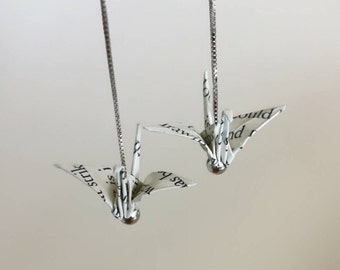 May include: Two origami paper cranes hanging from silver chains. The cranes are made from white paper with black text and are suspended from silver chains.