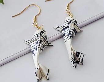 May include: A pair of origami-style fish earrings. The earrings are white with black musical note patterns and gold-coloured ear wires. The fish have detailed fins and tails, creating a unique and artistic accessory.