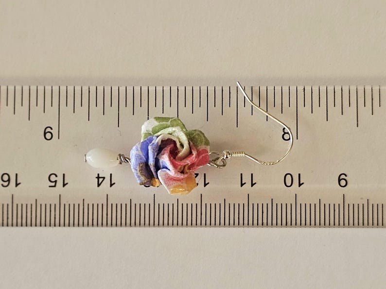 May include: A fabric rose earring with a white bead drop. The rose is made of pink, blue, yellow, and green fabric. The earring, including the hook, is approximately 3.2 cm long.