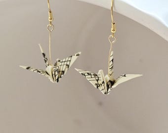 May include: A pair of gold-colored dangle earrings featuring origami crane pendants. The paper cranes are cream-colored with black musical notes printed on them. The earrings are suspended from gold-colored fish hook ear wires.